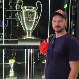 RoKa Renovation Robert Karpiński - Mężczyzna w czapce pozuje obok gabloty z trofeami sportowymi, w tym Pucharem Europy. Oświetlenie LED podkreśla eksponaty na czarnym tle.