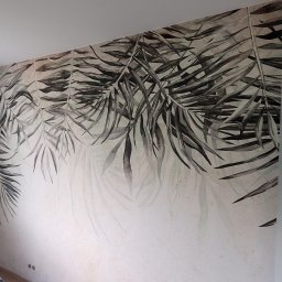 RoKa Renovation Robert Karpiński - Ściana z fototapetą w motywie liści palmowych, częściowo zasłonięta folią ochronną. Widoczne gniazdka elektryczne i fragment podłogi. Świeżo położona tapeta.