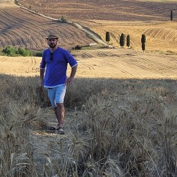 RoKa Renovation Robert Karpiński - Mężczyzna w kapeluszu stoi na polu zboża z toskańskim krajobrazem w tle. Widoczne cyprysy i pagórkowate tereny.