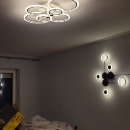 RoKa Renovation Robert Karpiński - Nowoczesna lampa sufitowa z okręgów i geometryczny kinkiet na szarej ścianie. Meble zabezpieczone folią malarską. Oświetlenie wnętrza po montażu.