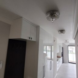 RoKa Renovation Robert Karpiński - Białe, błyszczące szafki w przedpokoju z kryształowymi lampami sufitowymi. Widok na kuchnię i okno. Nowoczesne wykończenie wnętrza.