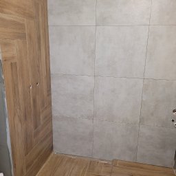 RoKa Renovation Robert Karpiński - Wykończona łazienka z płytkami imitującymi drewno na ścianie i podłodze, ułożonymi w jodełkę. Ściana wyłożona szarymi, kwadratowymi płytkami. Widoczne otwory na gniazdka.