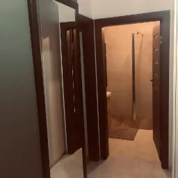 Widok z korytarza na wykończoną łazienkę z kabiną prysznicową typu walk-in i brązowymi drzwiami, widzianą przez otwarte drzwi.