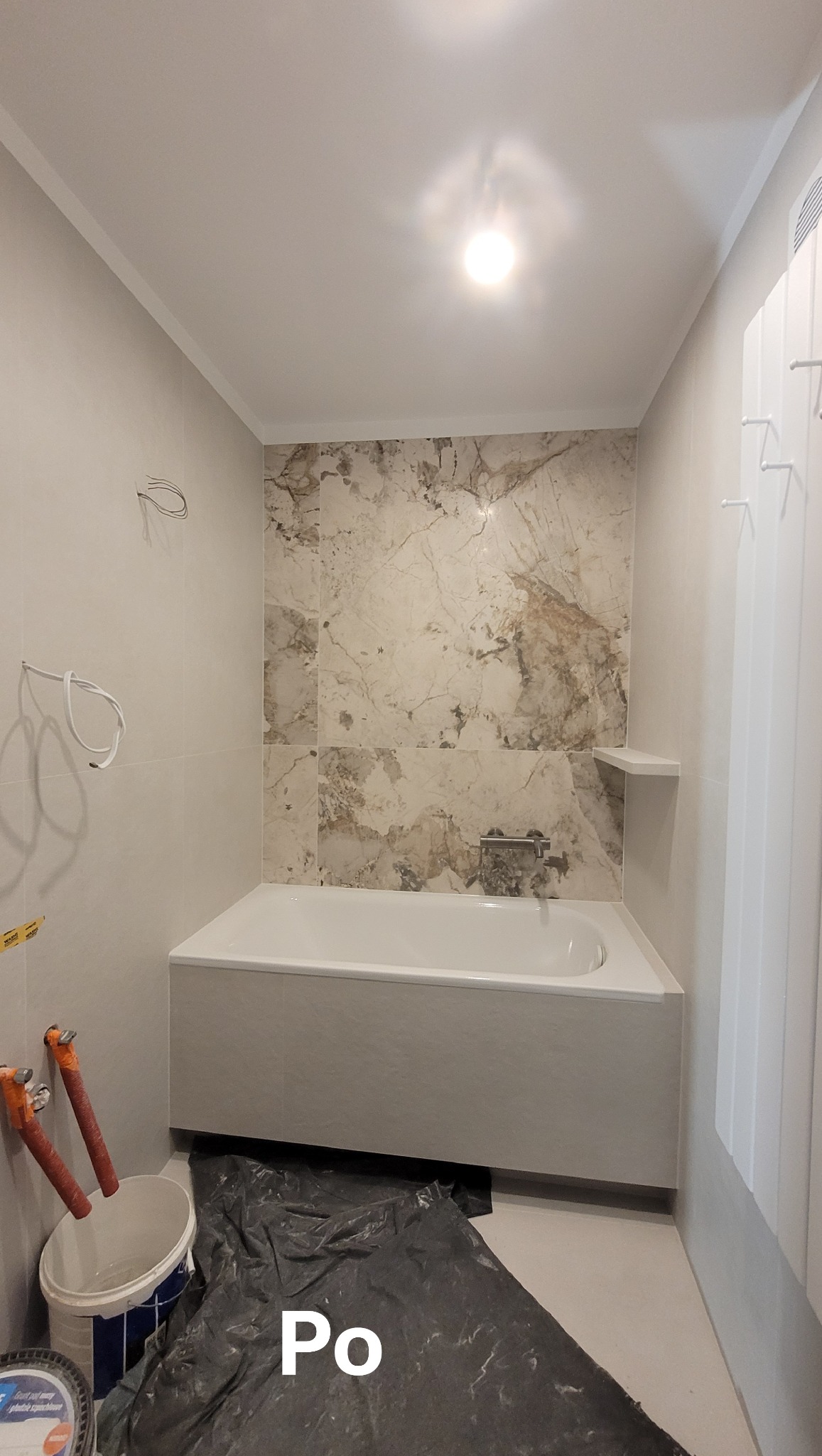 Wykończona łazienka z wanną w zabudowie, marmurowe płytki na ścianie, grzejnik drabinkowy. Widoczne przygotowania do dalszych prac.