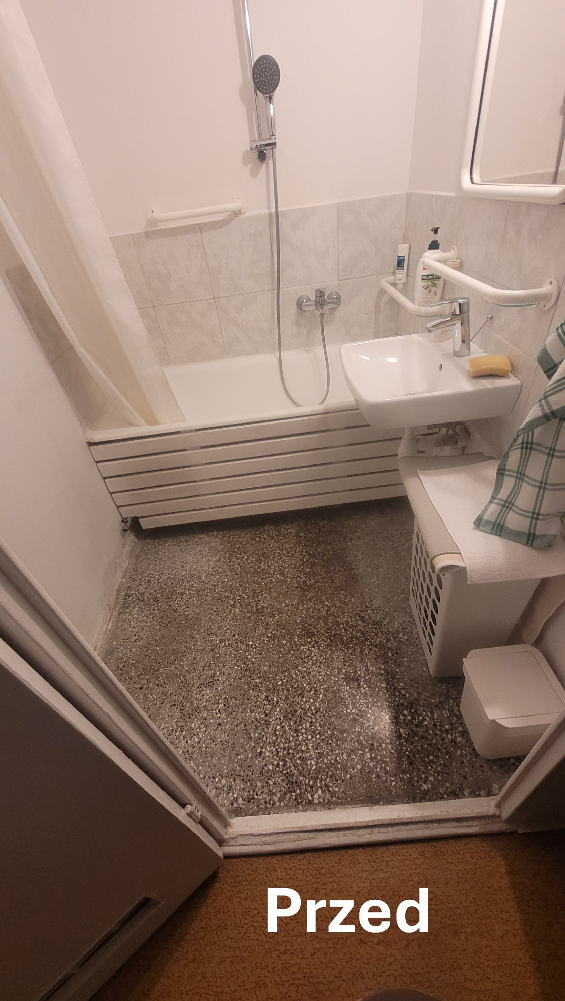 Łazienka z wanną, umywalką i grzejnikiem drabinkowym. Widoczne uchwyty dla niepełnosprawnych. Zdjęcie 'przed' remontem, z widoczną starą podłogą.