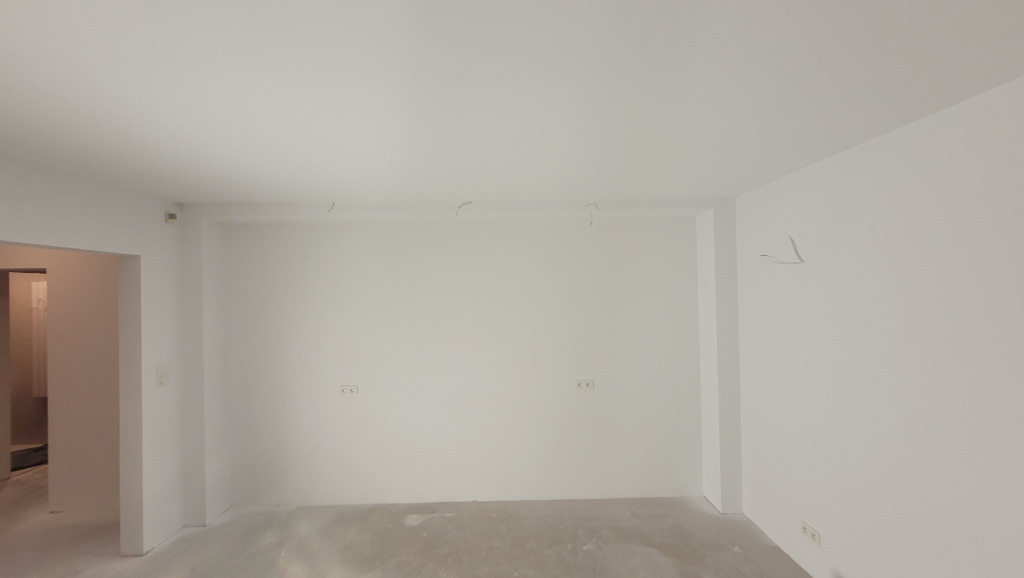 Wnętrze po malowaniu: białe ściany, widoczne gniazdka elektryczne i wystające kable ze ściany. Podłoga przygotowana do dalszych prac wykończeniowych.