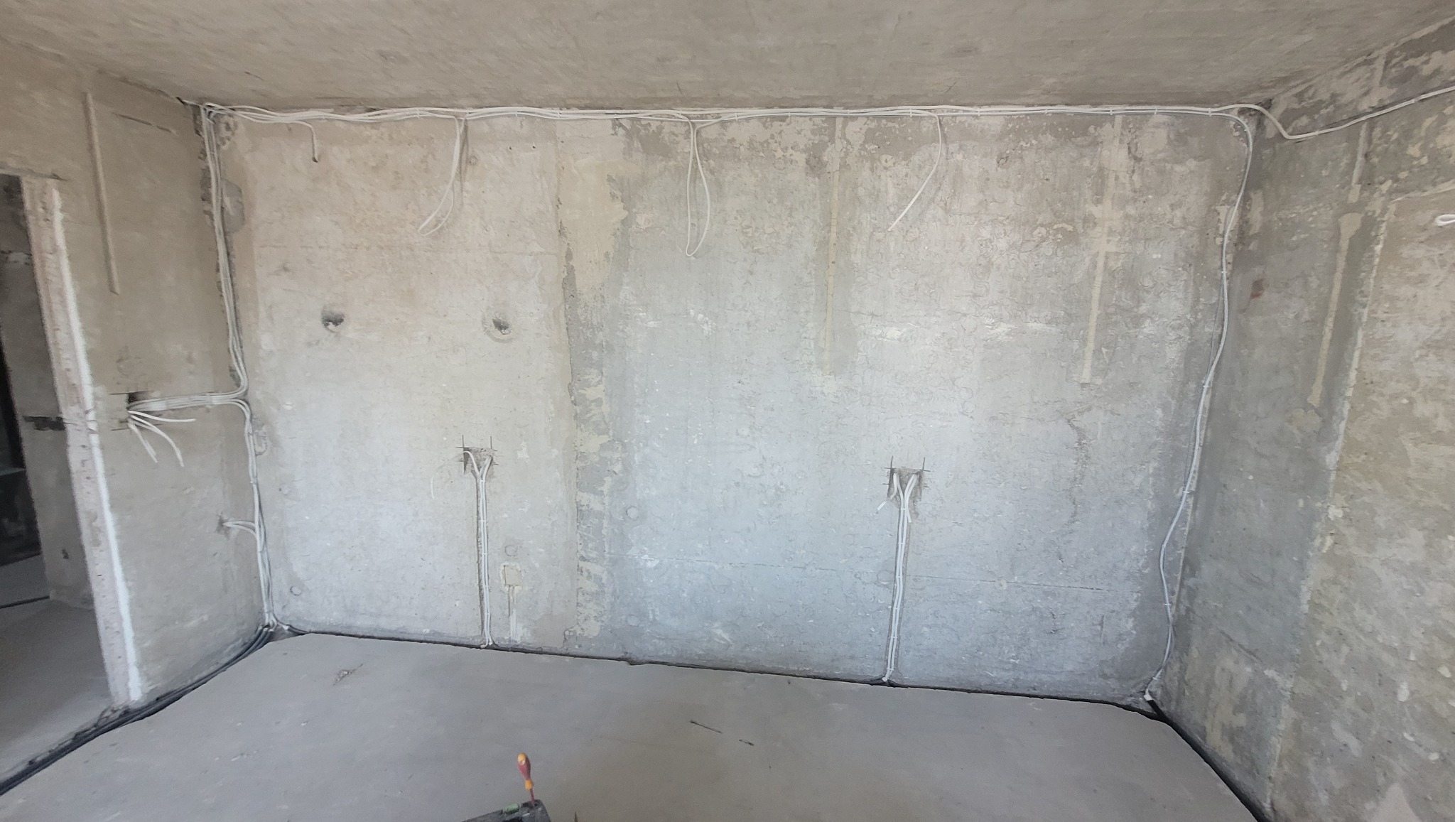 Surowy betonowy pokój w trakcie prac elektrycznych: widoczne kable prowadzone po ścianach i suficie, przygotowane punkty instalacyjne. Minimalistyczny, industrialny charakter.