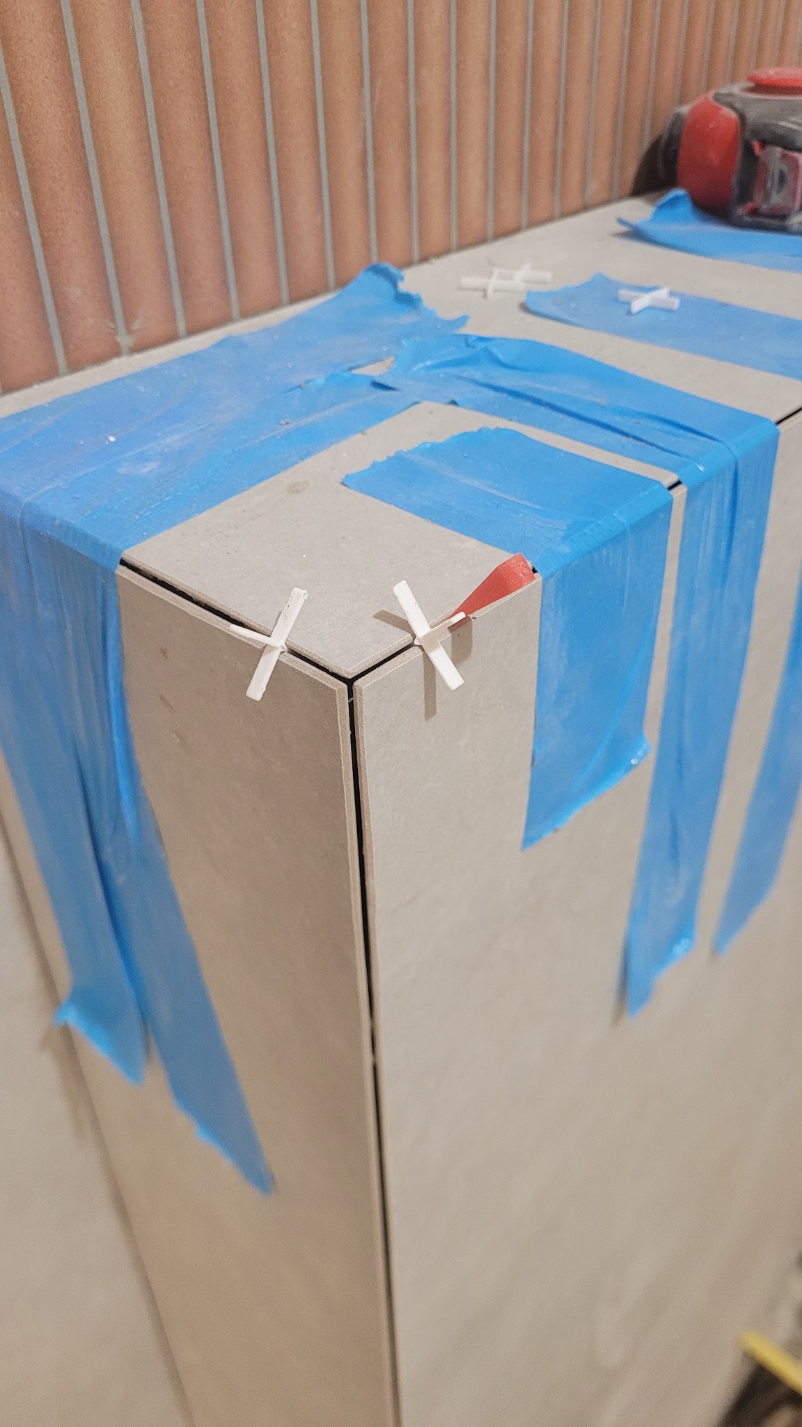 Układanie płytek na narożniku, zabezpieczone niebieską folią i krzyżykami dystansowymi. Widoczna ściana z cegieł.