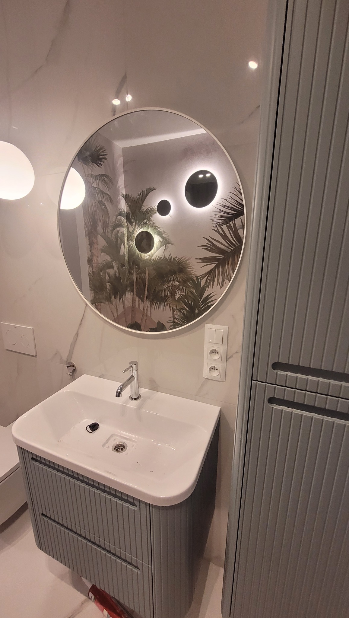 Nowoczesna łazienka z okrągłym lustrem z podświetleniem LED, umywalką w szafce z szarymi lamelami i marmurową ścianą. Widoczne gniazdka elektryczne.