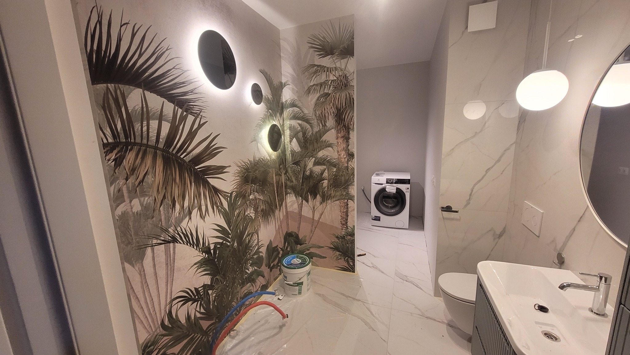 Wykończona łazienka z motywem tropikalnym na tapecie, okrągłe, podświetlane lampy na ścianie, białe płytki imitujące marmur i pralka w tle. Nowoczesny design.