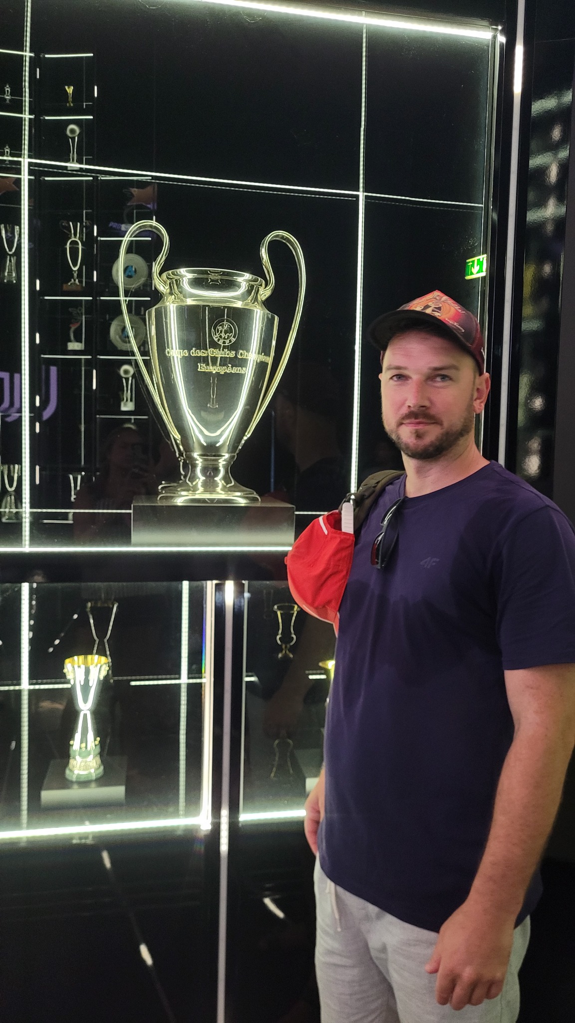 Mężczyzna w czapce pozuje obok gabloty z trofeami sportowymi, w tym Pucharem Europy. Oświetlenie LED podkreśla eksponaty na czarnym tle.