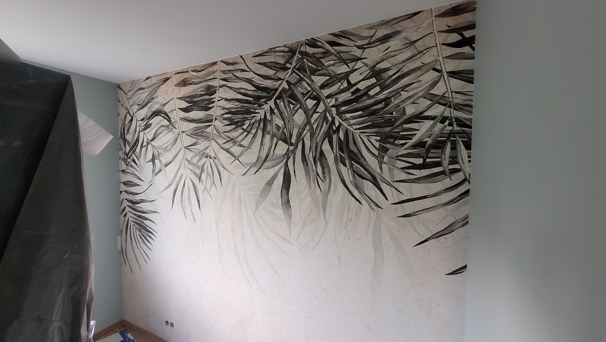 Ściana z fototapetą w motywie liści palmowych, częściowo zasłonięta folią ochronną. Widoczne gniazdka elektryczne i fragment podłogi. Świeżo położona tapeta.