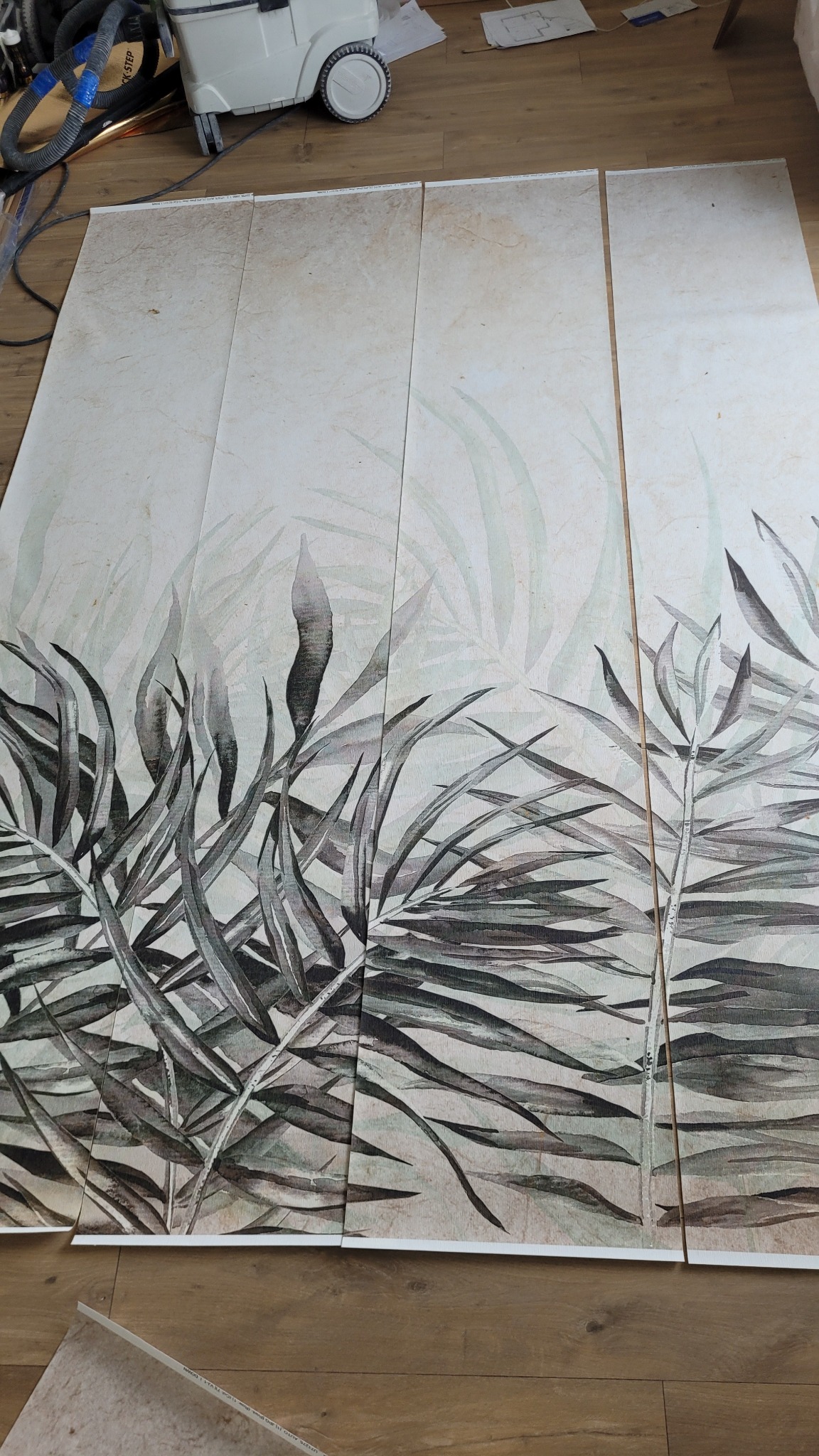 Cztery panele z motywem roślinnym, ułożone obok siebie na podłodze, tworzące spójny wzór. Widoczny odkurzacz przemysłowy w tle, sugerujący prace wykończeniowe.
