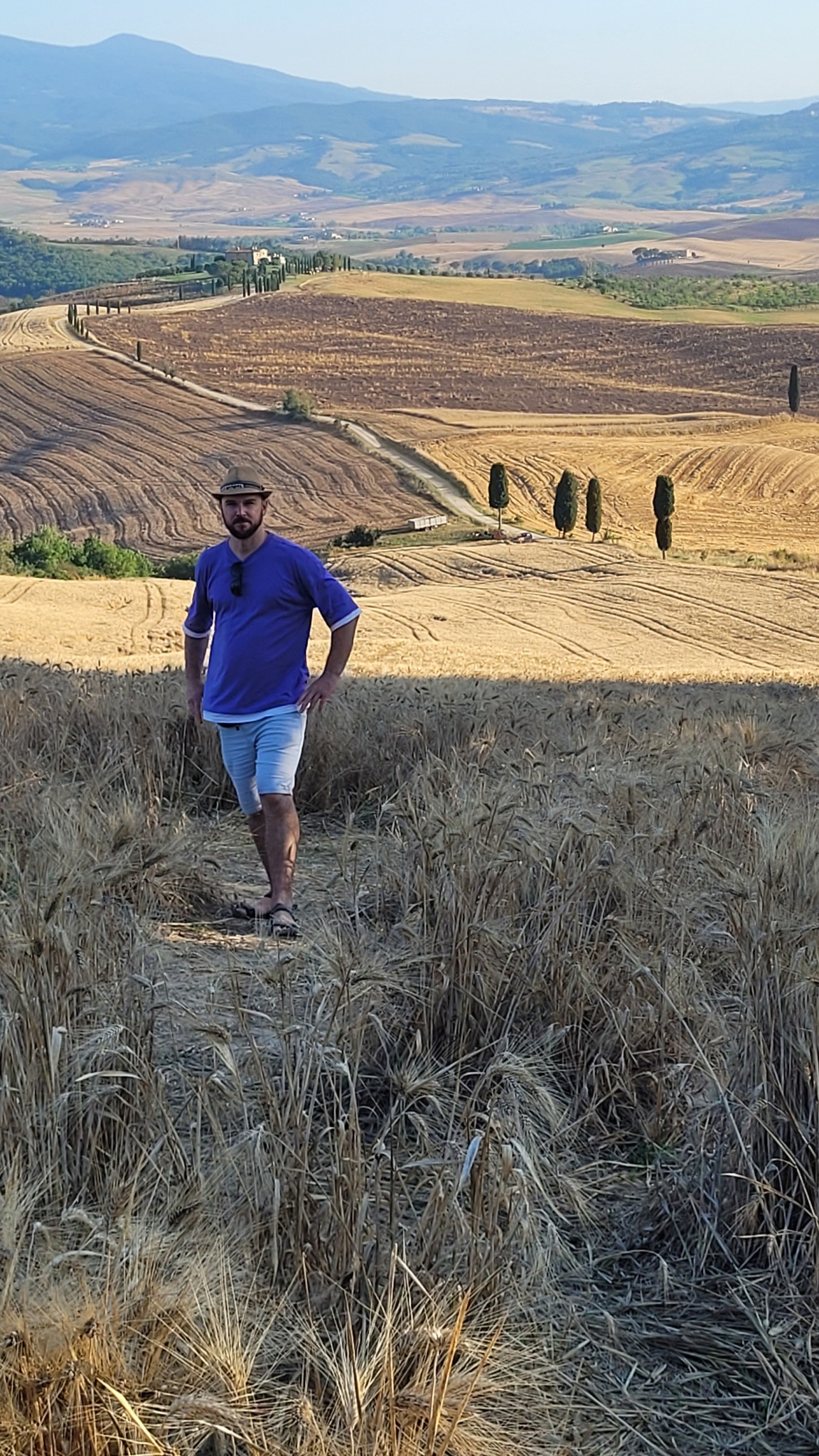 Mężczyzna w kapeluszu stoi na polu zboża z toskańskim krajobrazem w tle. Widoczne cyprysy i pagórkowate tereny.