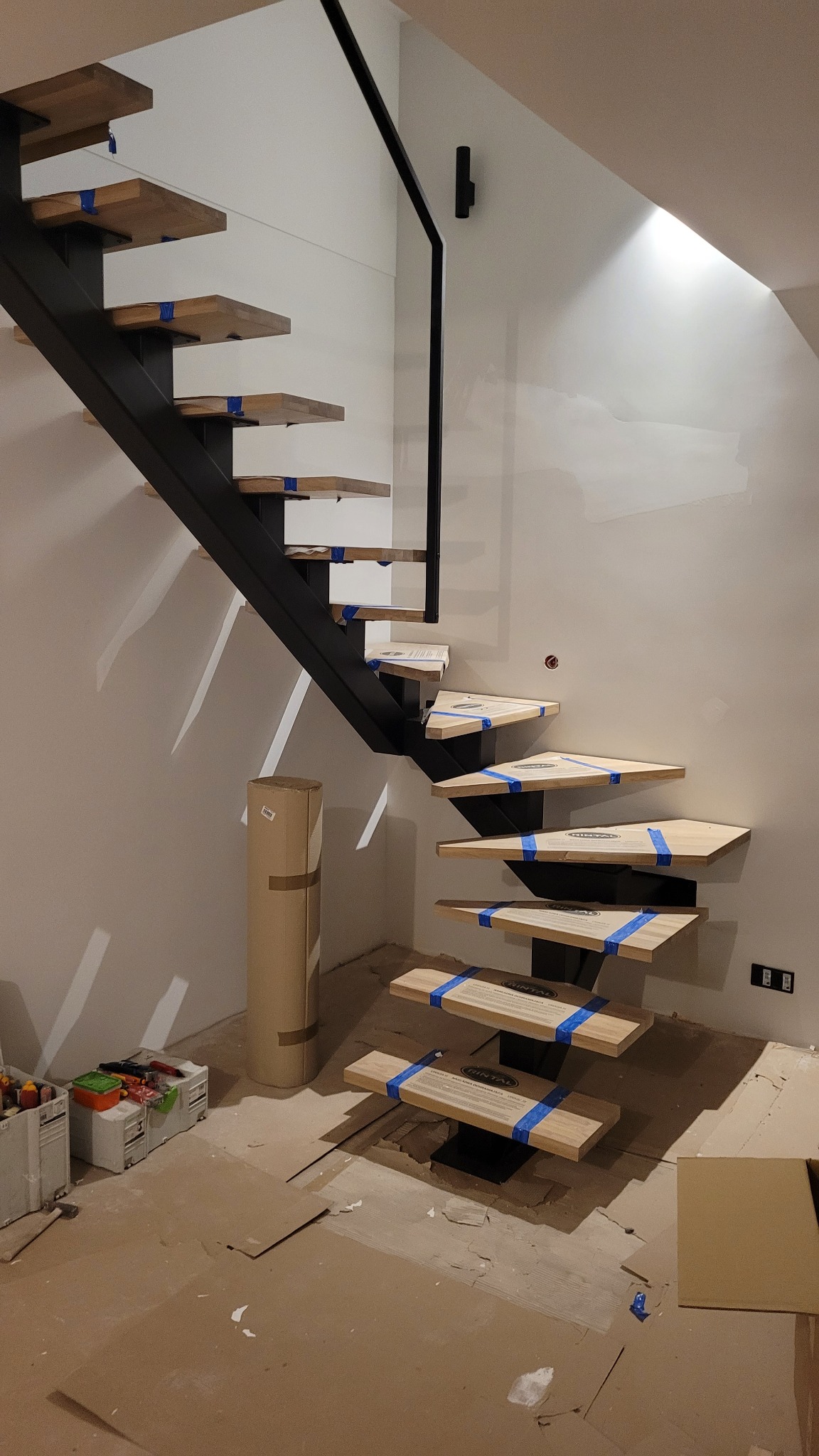 Nowoczesne schody wiszące z drewnianymi stopniami zabezpieczonymi niebieską taśmą, czarna metalowa konstrukcja, widoczne elementy wykończeniowe w trakcie montażu.