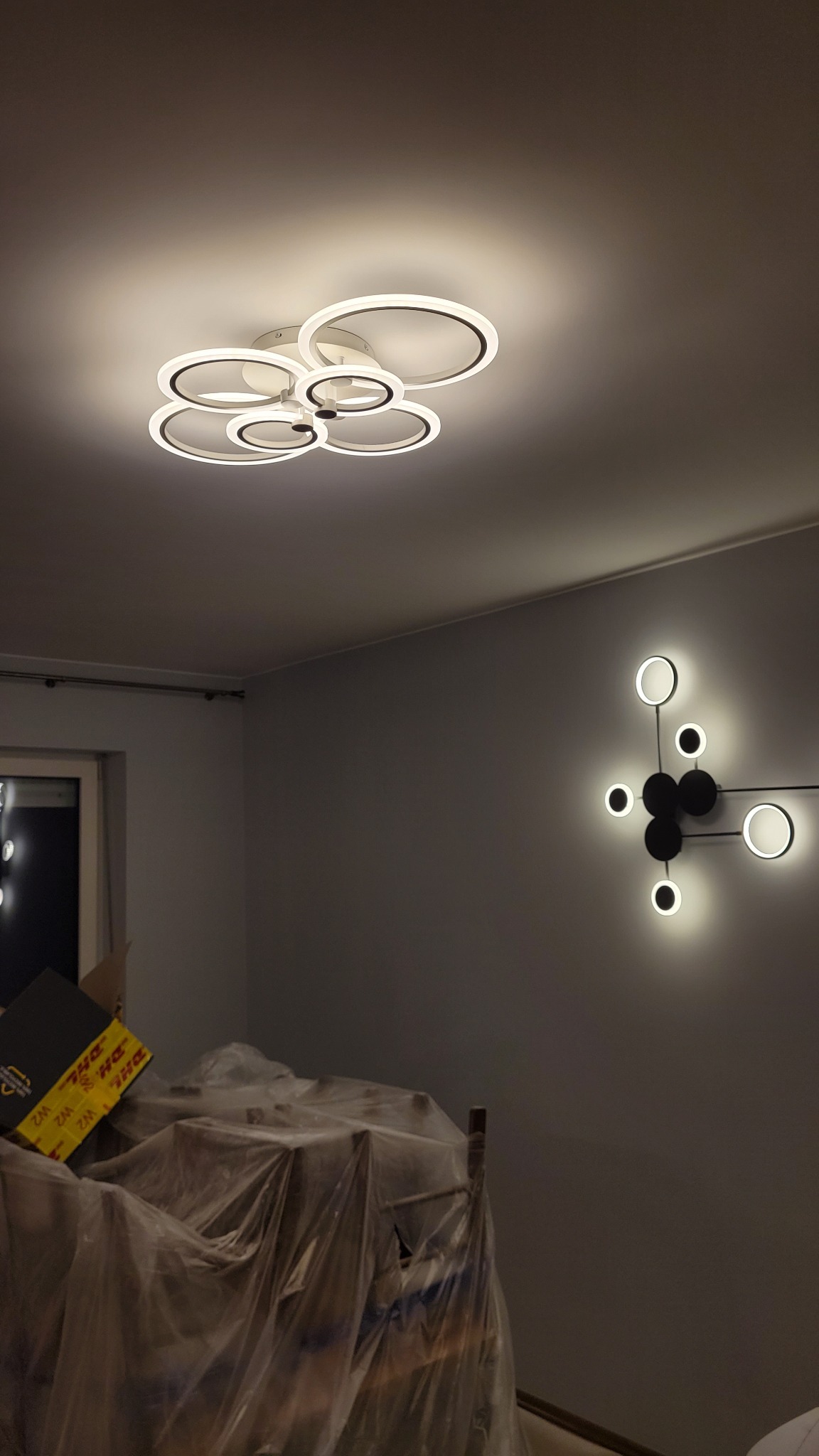 Nowoczesna lampa sufitowa z okręgów i geometryczny kinkiet na szarej ścianie. Meble zabezpieczone folią malarską. Oświetlenie wnętrza po montażu.