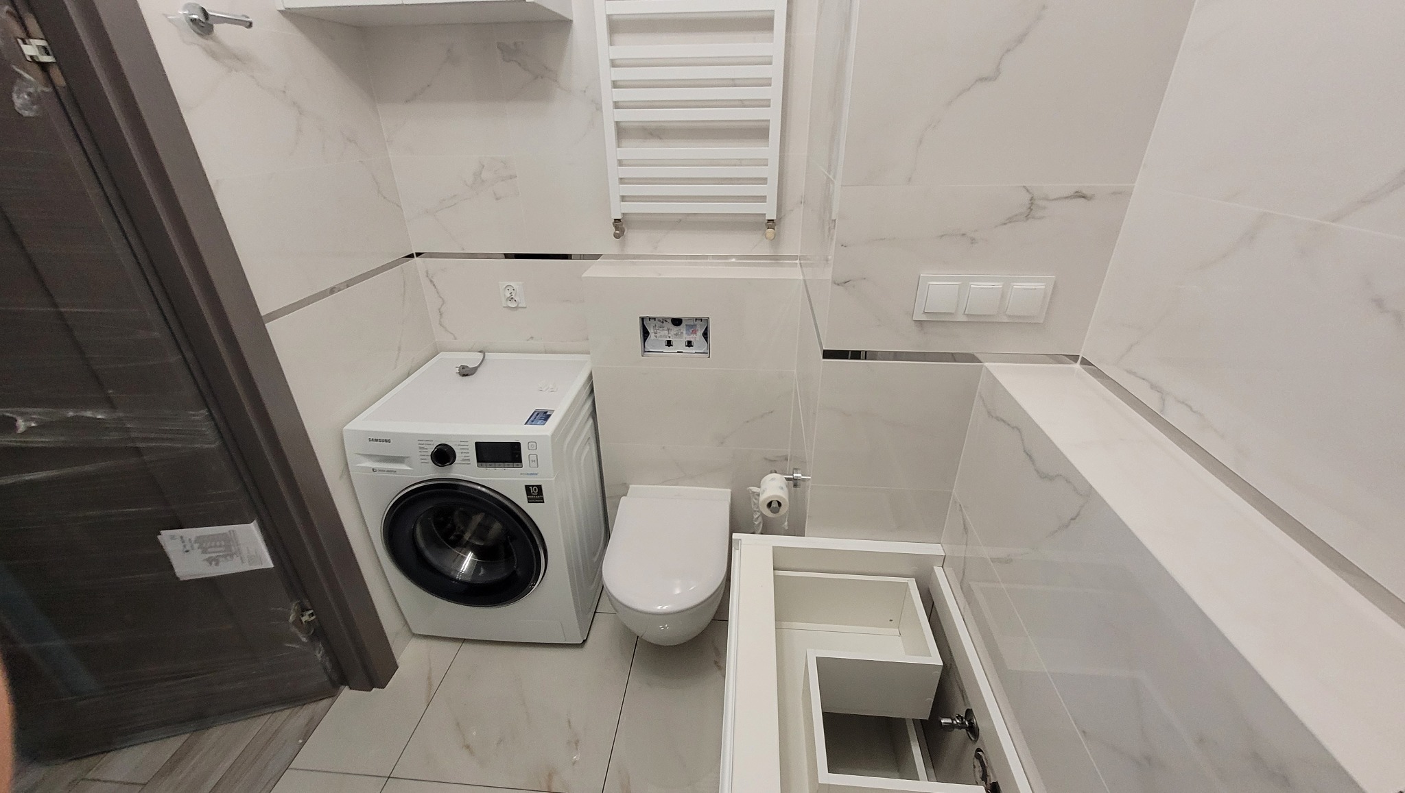 Nowoczesna łazienka z marmurowymi płytkami, pralką Samsung, podwieszaną toaletą i grzejnikiem drabinkowym. Minimalistyczny design z akcentami chromu.