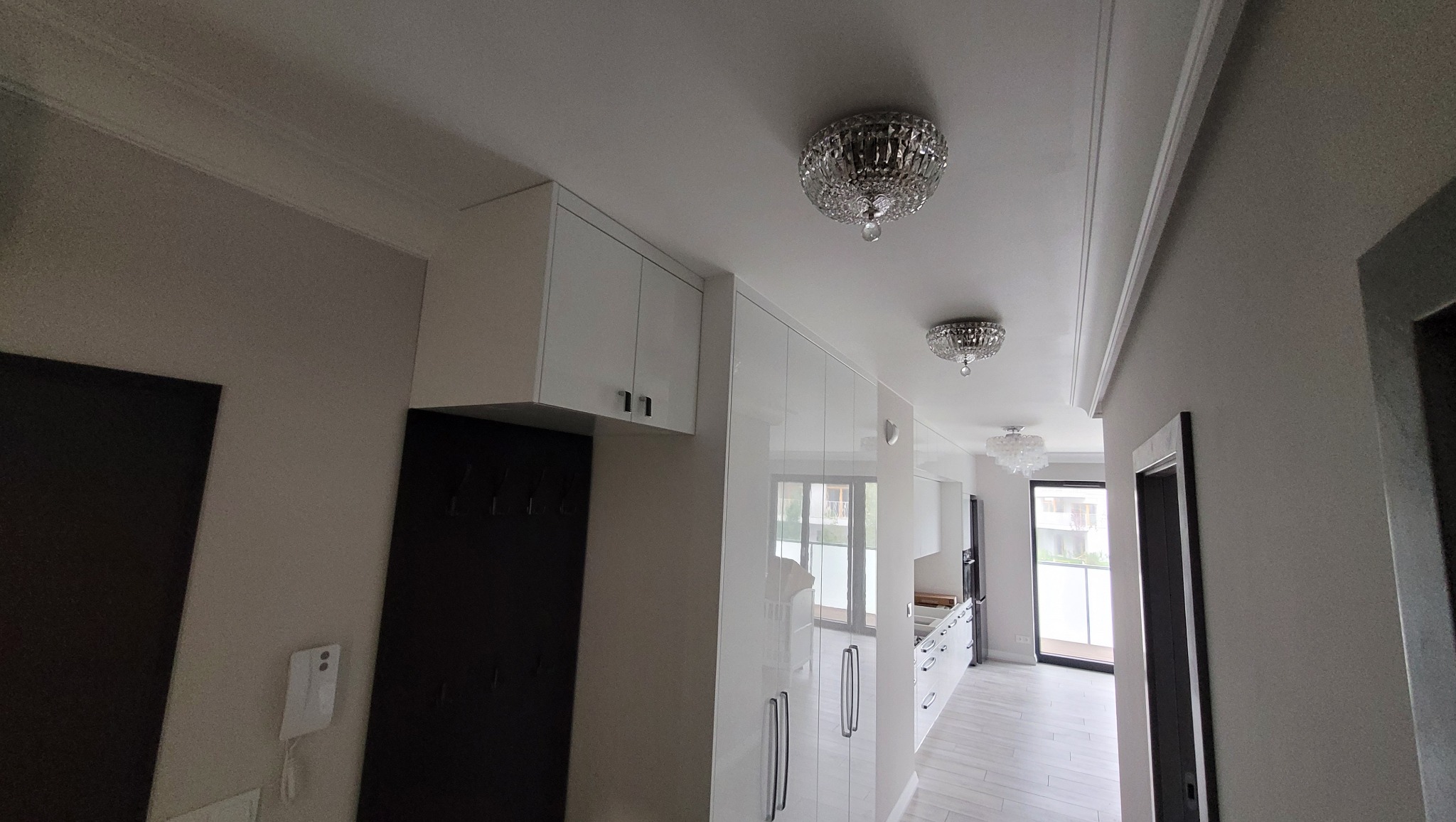 Białe, błyszczące szafki w przedpokoju z kryształowymi lampami sufitowymi. Widok na kuchnię i okno. Nowoczesne wykończenie wnętrza.