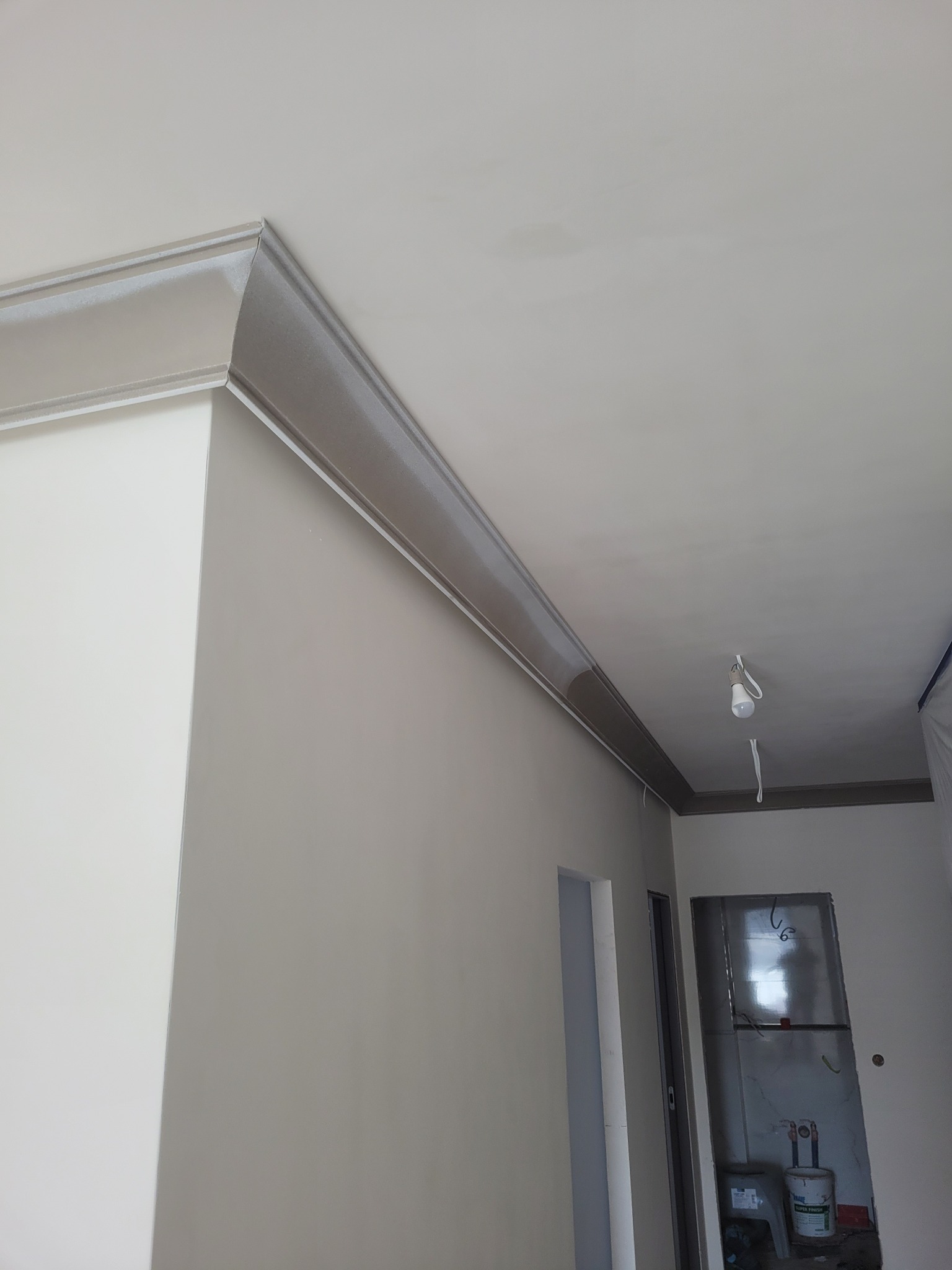 Wykończone wnętrze z listwami dekoracyjnymi przy suficie, pomalowane ściany na jasny kolor. Widoczne drzwi i fragment pomieszczenia w trakcie prac wykończeniowych.