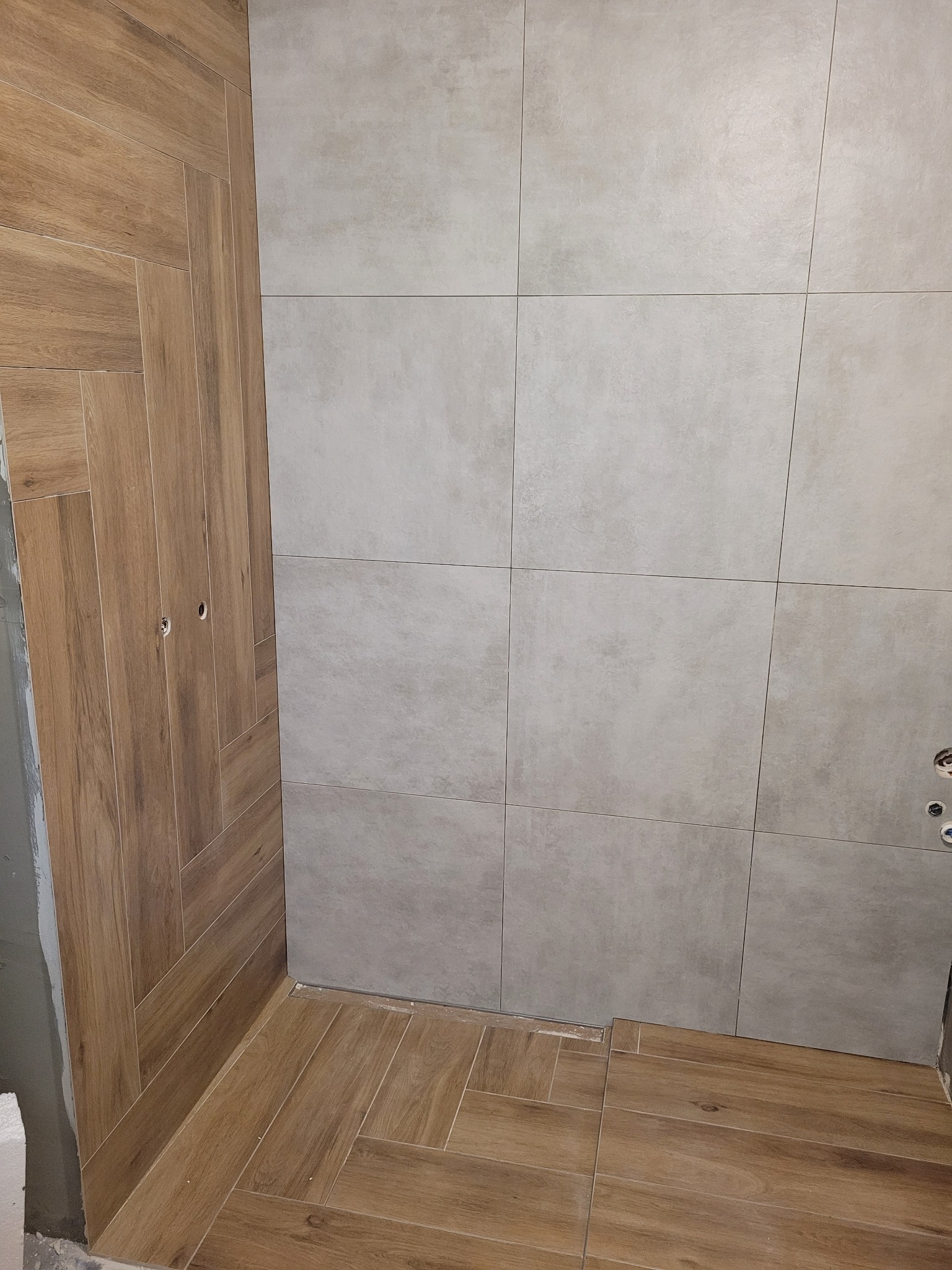 Wykończona łazienka z płytkami imitującymi drewno na ścianie i podłodze, ułożonymi w jodełkę. Ściana wyłożona szarymi, kwadratowymi płytkami. Widoczne otwory na gniazdka.