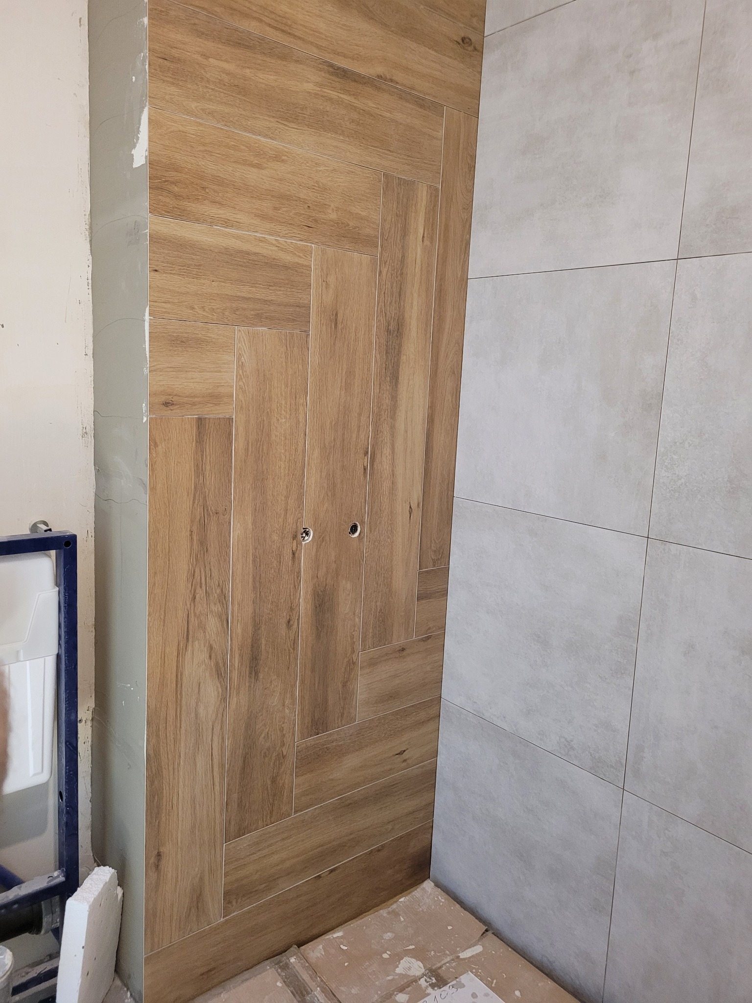 Ułożone płytki imitujące drewno w jodełkę na ścianie, obok szare płytki. Widoczne otwory na armaturę. Fragment instalacji i surowa ściana w tle.