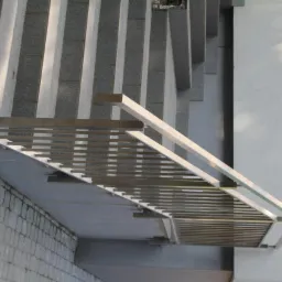 Zewnętrzna balustrada nierdzewna z poziomymi prętami, zamontowana przy schodach wejściowych do budynku, widoczna z boku, z fragmentem tablicy informacyjnej na ścianie.