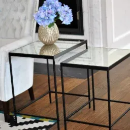 Dwa stoliki kawowe z czarnymi, minimalistycznymi ramami i szklanymi blatami, na jednym wazon z niebieskimi hortensjami, obok fotel z jasną tapicerką i fragment wzorzystego dywanu.