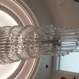 Kaskadowy żyrandol z przezroczystych, szklanych rurek zawieszony na metalowej konstrukcji, widok z dołu na tle okrągłego, podświetlanego sufitu z dekoracyjnymi listwami.