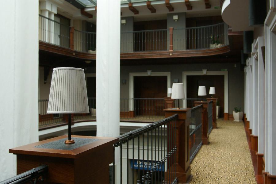 Wnętrze hotelu z metalową balustradą, kolumnami i lampami stołowymi z plisowanymi abażurami na drewnianych postumentach.
