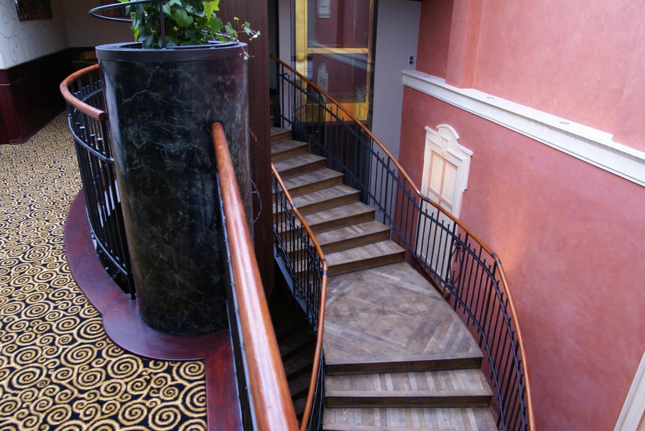 Zakręcone schody z drewnianymi stopniami, czarną metalową balustradą i drewnianą poręczą, obok marmurowego filaru z rośliną, na tle wzorzystej podłogi i ściany w odcieniach różu.
