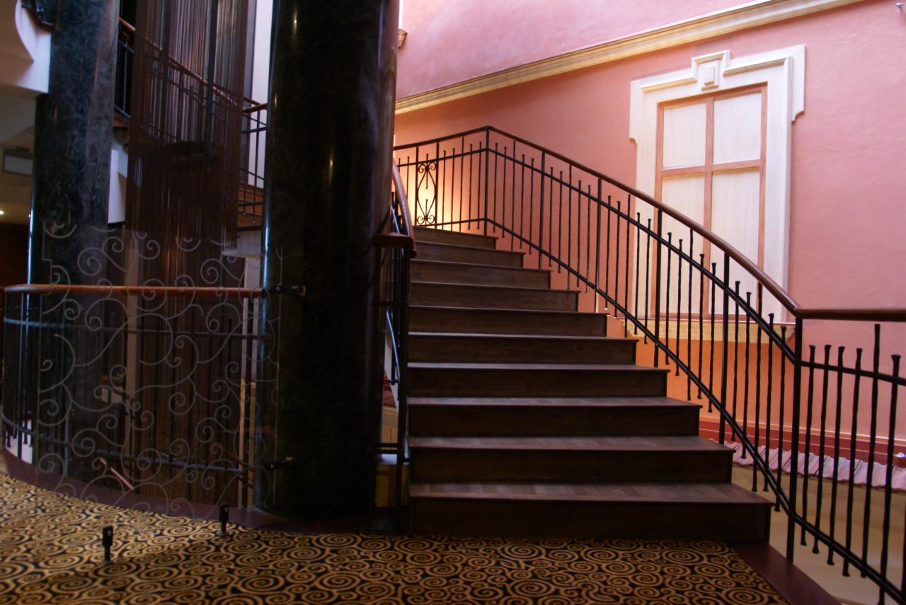 Klatka schodowa z drewnianymi stopniami i czarną, kutą balustradą w budynku o różowych ścianach i marmurowych kolumnach.