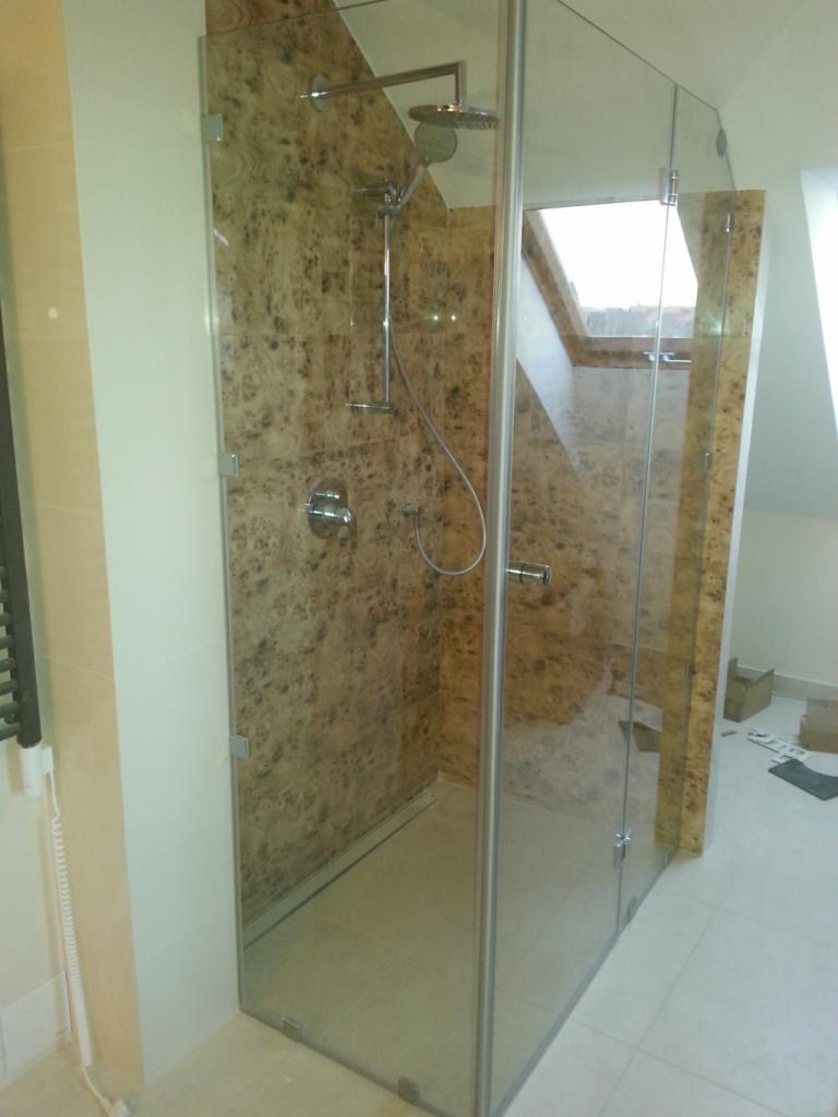 Kabina prysznicowa ze szklanymi ścianami i drzwiami, z widocznym brodzikiem i baterią, wykończona płytkami imitującymi drewno na ścianie w tle, w nowoczesnej łazience z oknem dachowym.