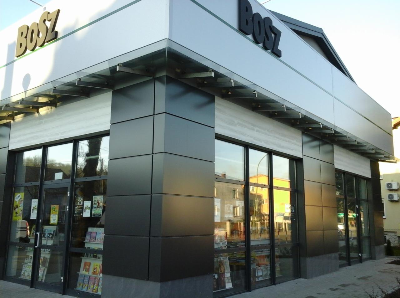 Nowoczesna fasada budynku z dużymi, przeszklonymi witrynami i szklanym daszkiem, wykończona panelami w odcieniach szarości i czerni.