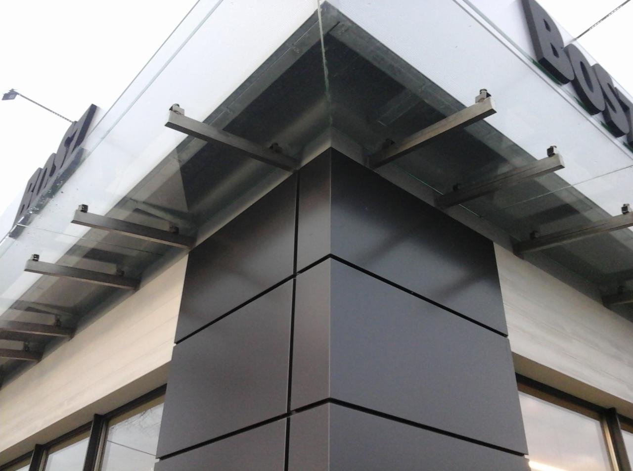 Szklany daszek nad wejściem do budynku, wsparty na metalowych wspornikach, z widocznymi mocowaniami do szklanej tafli i elewacji.