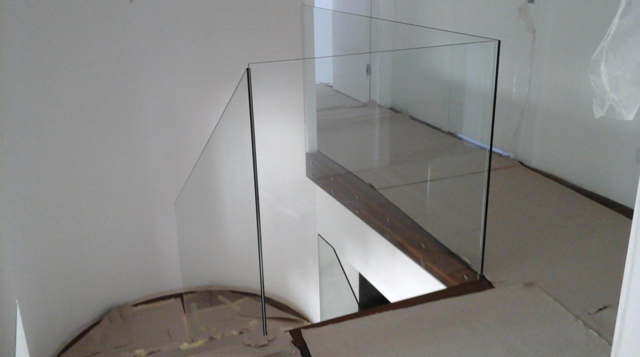 Szklana barierka zabezpieczająca schody, z drewnianymi elementami mocującymi, widok z góry, białe ściany w tle.