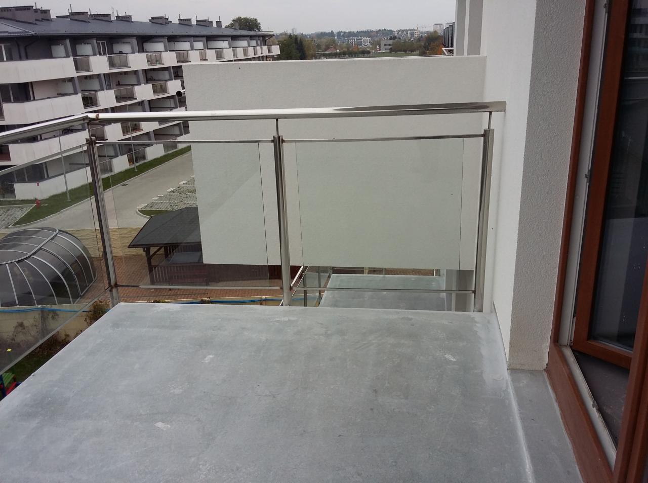 Balkon z nowoczesną, szklaną balustradą z elementami ze stali nierdzewnej, widok z góry na szarą, betonową podłogę balkonu i otoczenie budynku.
