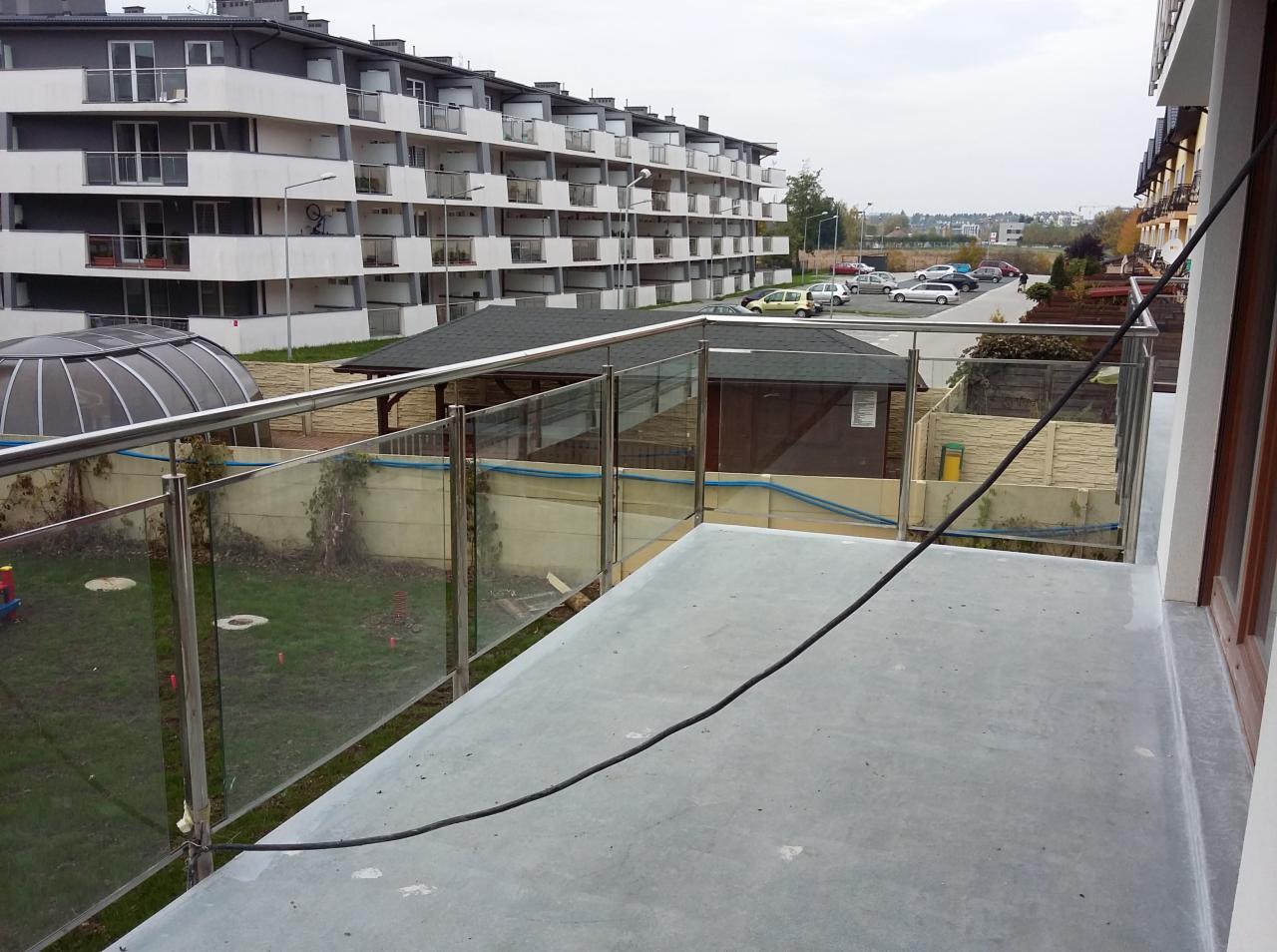Szklana balustrada balkonowa z nierdzewnymi słupkami, widok z góry na betonową podłogę balkonu, w tle zabudowa wielorodzinna i parking.