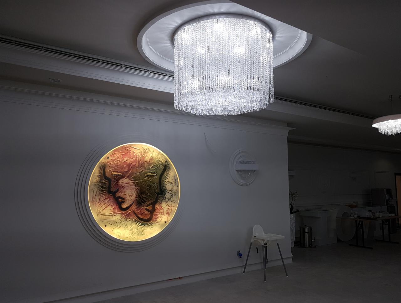 Kryształowy żyrandol kaskadowy zawieszony pod sufitem, okrągła, podświetlana dekoracja ścienna z motywem twarzy, białe ściany z dekoracyjnymi listwami, w tle widoczny drugi żyrandol.