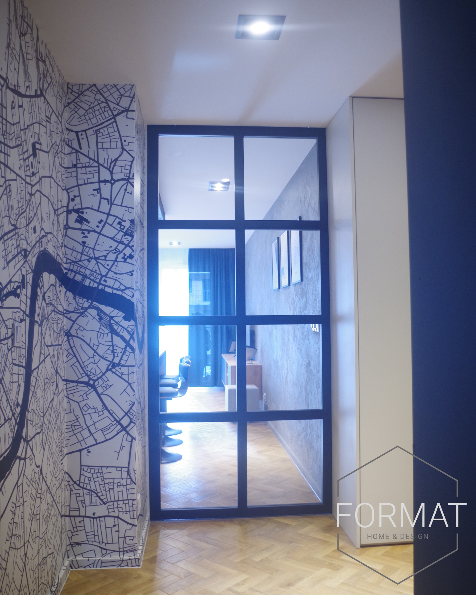 Korytarz z tapetą w mapę miasta i czarnymi, przeszklonymi drzwiami do salonu z parkietem w jodełkę, widoczne trzy obrazy na ścianie, logo firmy Format Home & Design w prawym dolnym rogu.