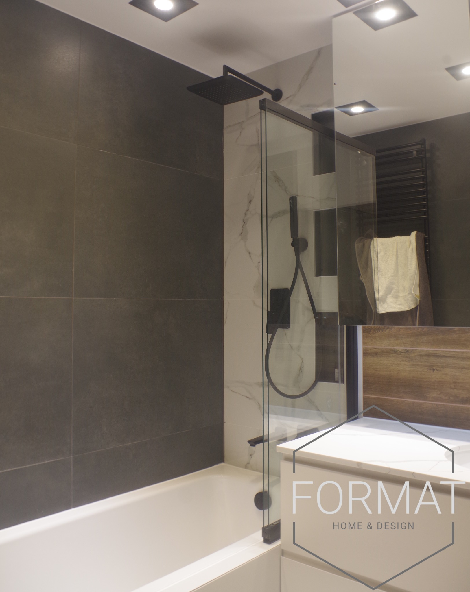 Nowoczesna łazienka z czarną armaturą, szklanym panelem prysznicowym i dużymi szarymi płytkami. Widoczny biały blat umywalki z logo firmy na pierwszym planie.