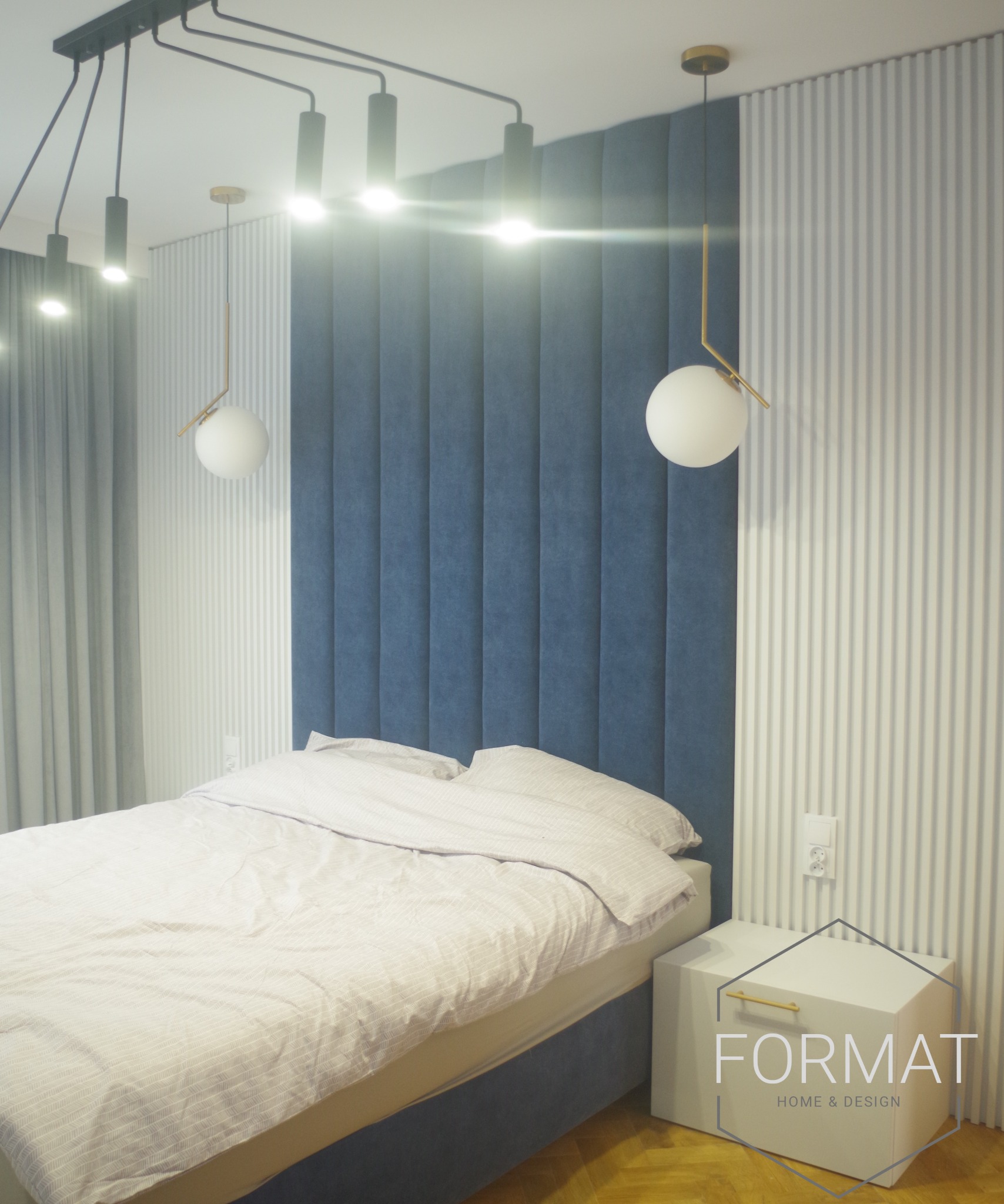 Nowoczesna sypialnia z łóżkiem z niebieskim zagłówkiem, nocną szafką z logo firmy, oraz designerskim oświetleniem sufitowym i wiszącymi lampami kulami.