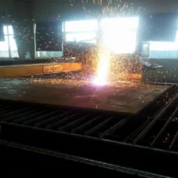 Automatyczne cięcie plazmowe grubej, ciemnej blachy stalowej na ruszcie, iskry i jasne światło w hali produkcyjnej z oknami.