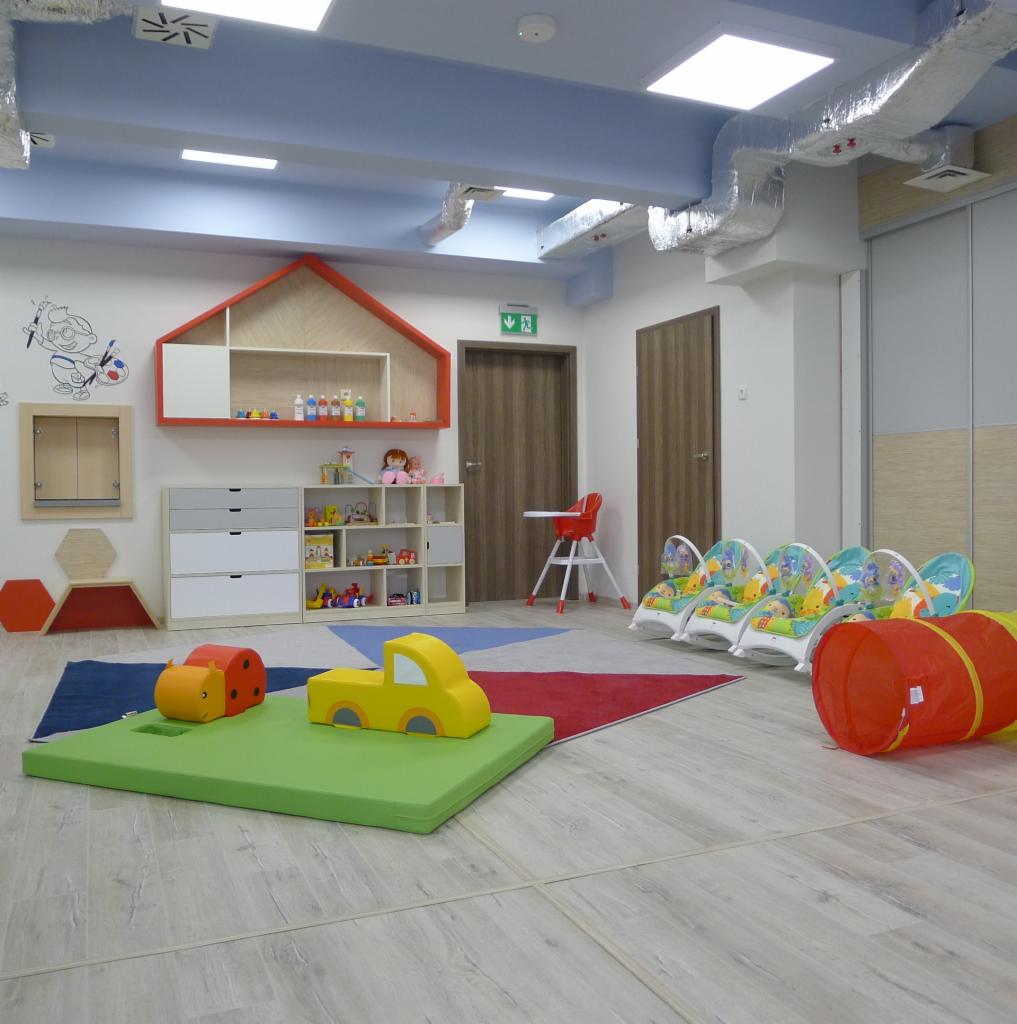 Jasna sala żłobka z drewnianą podłogą, wyposażona w kolorowe meble i zabawki, w tym żółty samochód i pomarańczowego hipopotama na zielonej macie, oraz tunel i leżaczki dla niemowląt.