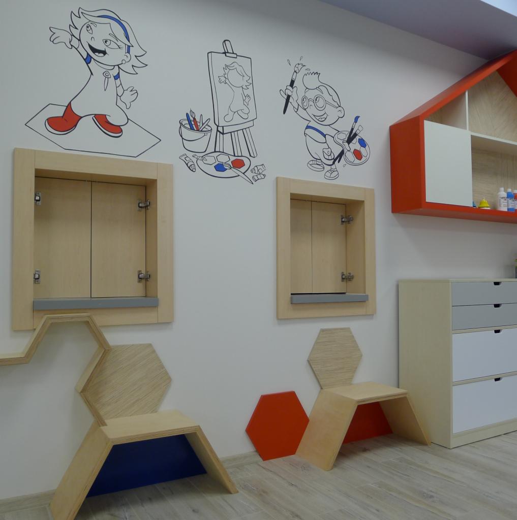 Jasna sala żłobka z meblami na wymiar: niskie szafki zamykane na klucz w ścianie, krzesełka w kształcie plastrów miodu i komoda z szufladami. Na ścianach rysunki dzieci i przybory malarskie.
