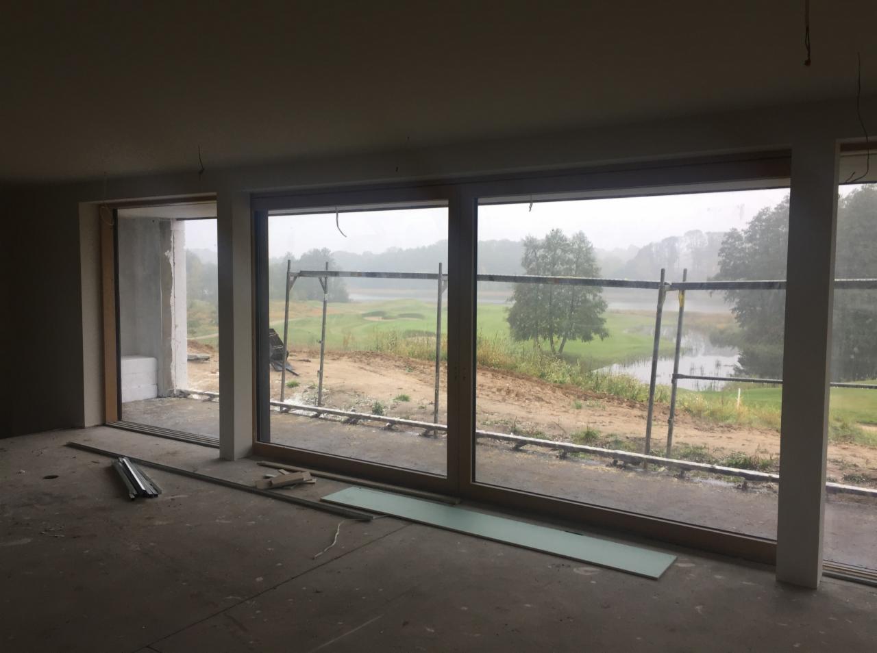 Surowy stan wnętrza z dużymi oknami z widokiem na pole golfowe w mglisty dzień. Widoczne profile okienne, płyty kartonowo-gipsowe i rusztowanie na zewnątrz. Wnętrze w trakcie wykończenia.