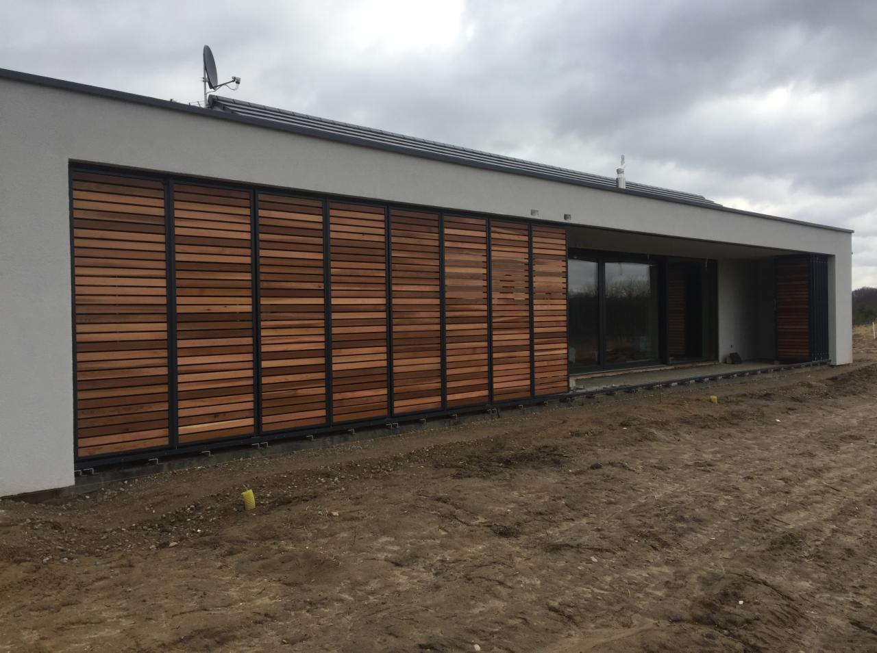 Nowoczesny dom jednorodzinny z drewnianymi panelami żaluzjowymi na fasadzie, zamontowanymi na metalowej ramie, przesłaniającymi okna i drzwi przesuwne, widoczne elementy instalacji satelitarnej...