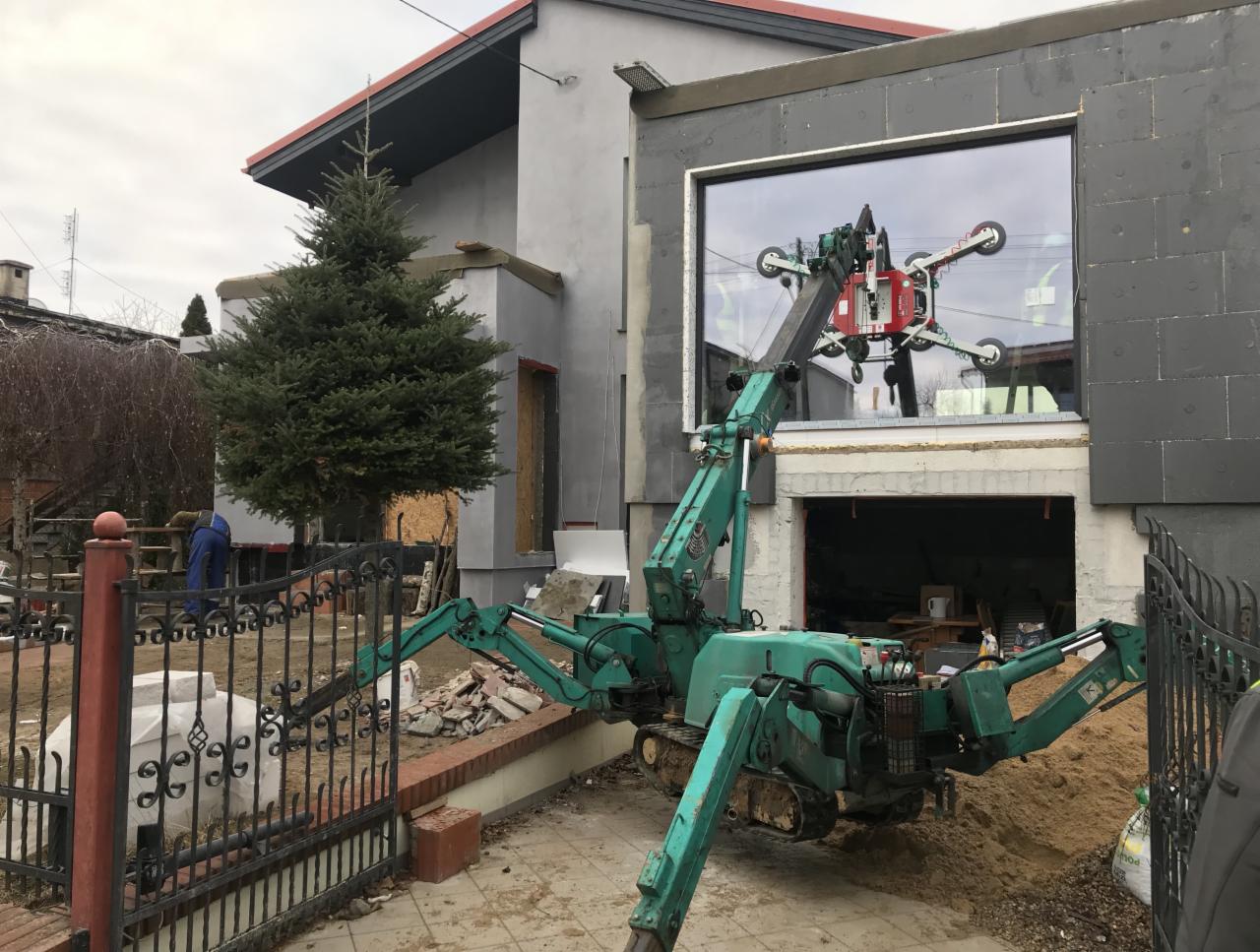 Montaż dużego okna w nowym budynku z użyciem specjalistycznego dźwigu z przyssawkami, widoczna elewacja w trakcie ocieplania, teren budowy.