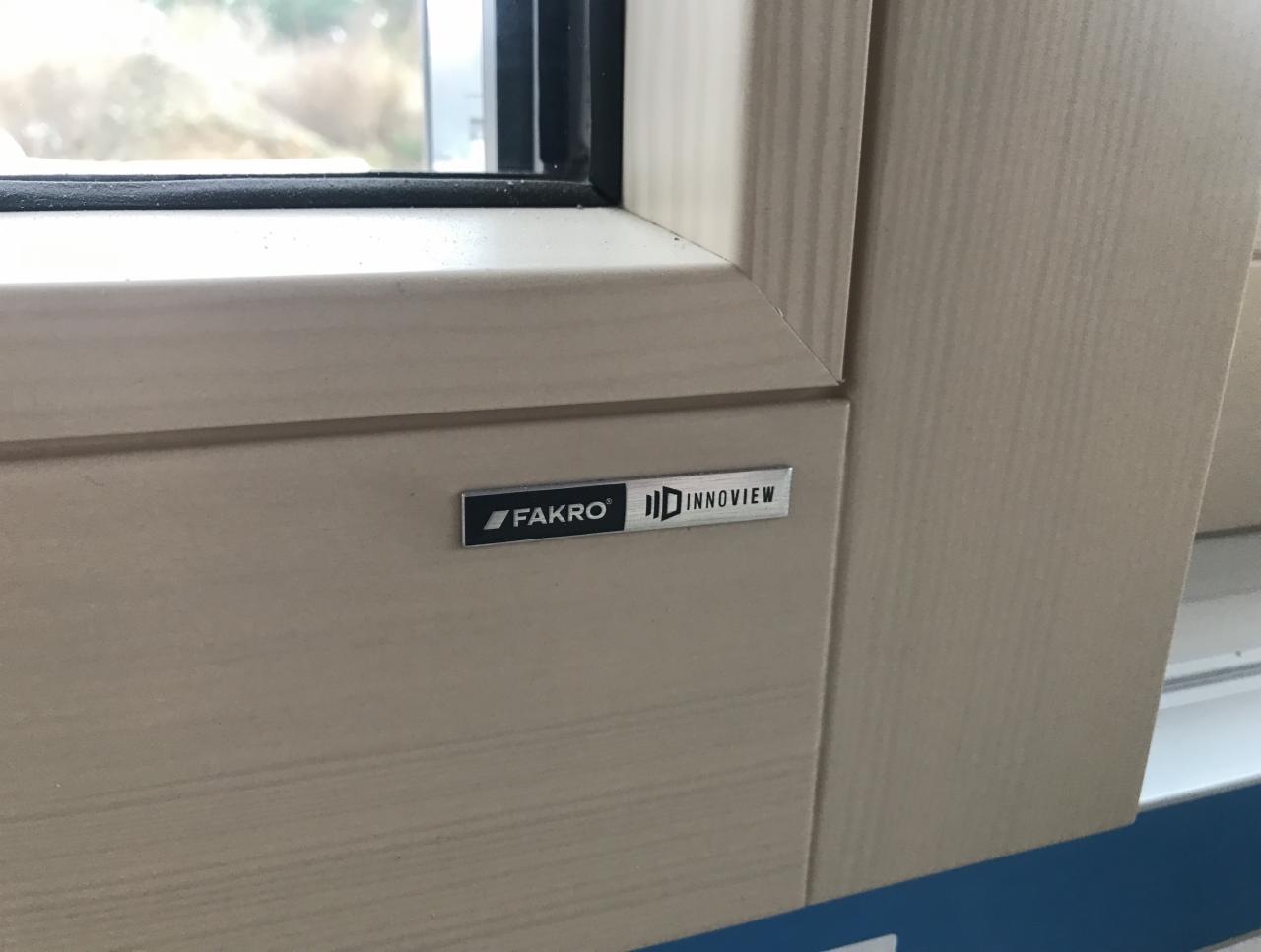 Fragment ramy okiennej z drewna w jasnym odcieniu z widoczną metalową tabliczką znamionową producenta Fakro Innoview.