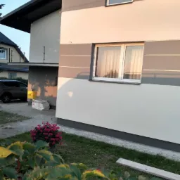 Dwukondygnacyjny dom z elewacją w odcieniach szarości i bieli, widok z niskiej perspektywy, z przodu roślinność i fragment chodnika.