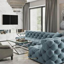 Justyna Tatys Luxury Interiors - Luksusowy salon z pikowaną sofą w odcieniach błękitu, szarym fotelem typu uszak, marmurową ścianą z telewizorem, kryształowym żyrandolem i portretem królowej z gumą balonową.